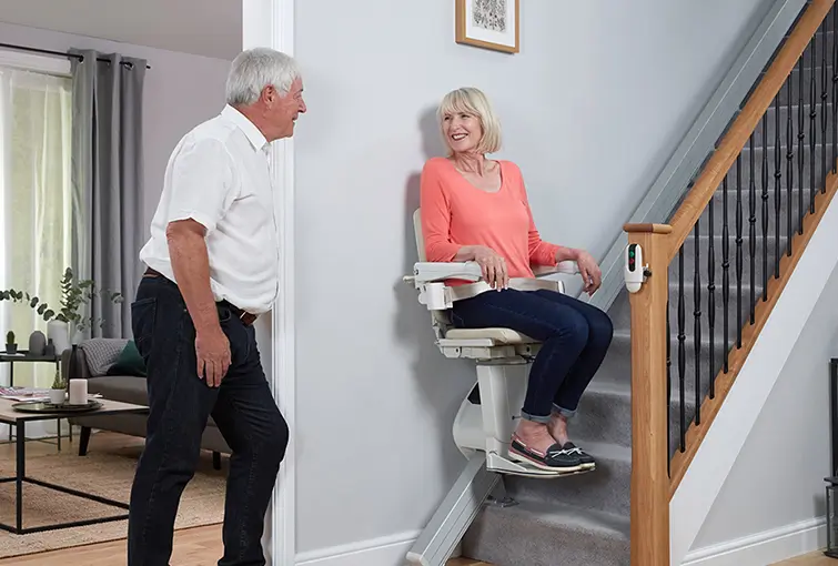 An elderly woman talks to her husband and uses her newly installed straight Handicare 1100 which is at the bottom of the stairs.