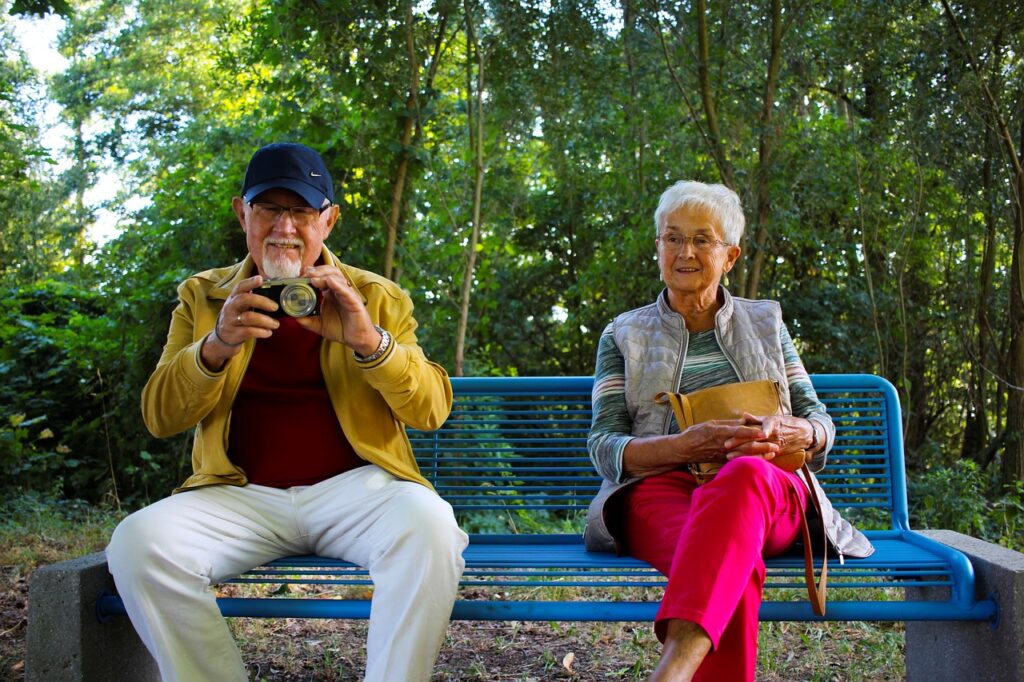 Elderly Couple sat in a park. One is taking a photo and taking part in a hobby and the other is relaxing.