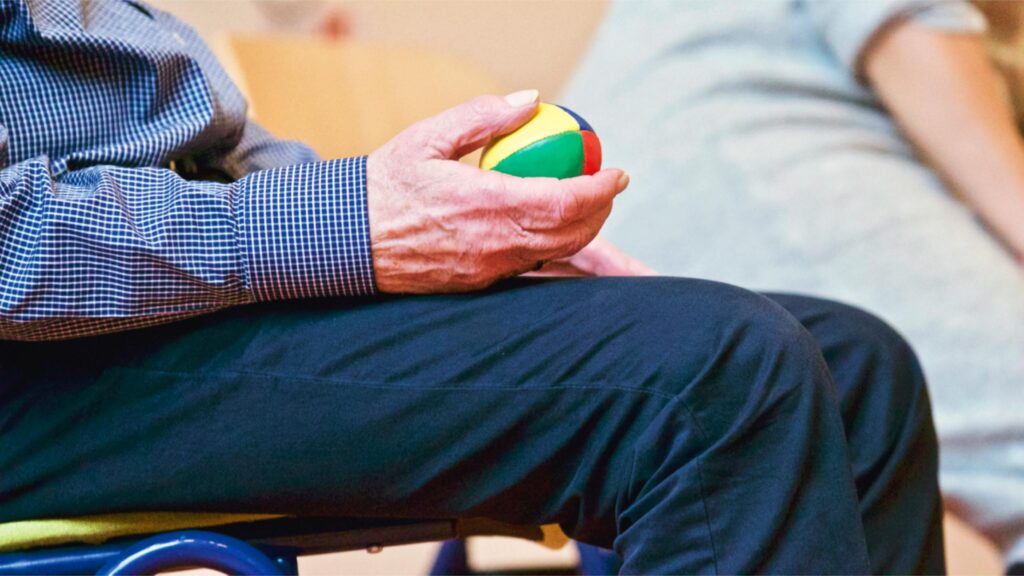 An Elderly man, a senior citizen holding a colourful ball whilst attending occupational therapy.